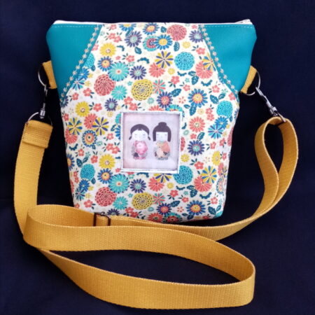 Le mini geishas.Sac bandoulière de forme presque carrée. Corps du sac en simili cuir turquoise. Devant ajout de coton à motif fleuri et d'un petit écusson avec de petites geishas d'import japonais. Sangle en coton moutarde pailletée et réglable. Une poche derrière fermée par un bouton, une poche dedans. Cherchez le Chas.