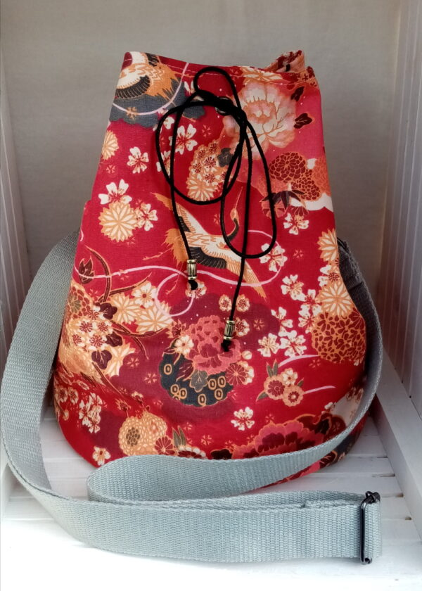 Sac sceau base ronde. Extérieur tissu renforcé en coton à motif japonais grues et fleurs sur fond rouge. Intérieur coton à petites fleurs. Sangle grise réglable. Une mini pochette assortie. Fermeture par un cordon noire passant par des œillets. Fond du sac renforcé semi rigide. Cherchez le Chas.